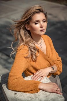 A middle-aged woman looks like a good blonde with curly beautiful hair and makeup on the background of the building. She is wearing a yellow sweater