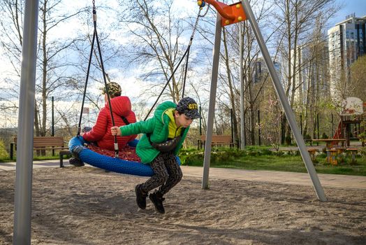 Two little kid boys having fun with swing on outdoor playground. Children, best friends and siblings swinging on warm sunny spring or autumn day. Active leisure with kids. Casual boy fashion.
