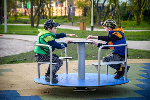 Brightly dressed twin brothers play on a carousel in an autumn park amid fallen yellow leaves. They are two years old.