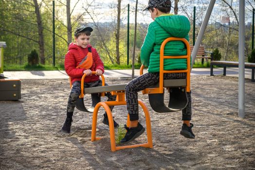 Two little kid boys having fun with swing on outdoor playground. Children, best friends and siblings swinging on warm sunny spring or autumn day. Active leisure with kids. Casual boy fashion.