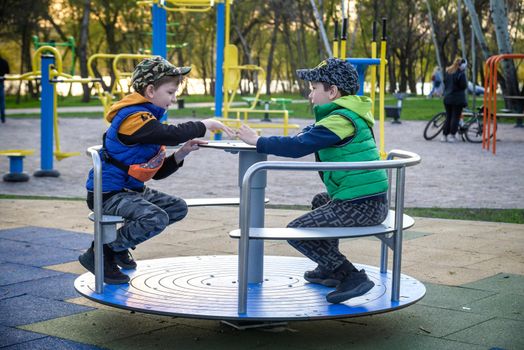 Brightly dressed twin brothers play on a carousel in an autumn park amid fallen yellow leaves. They are two years old.