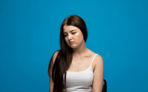 Portrait of upset young beautifil brunette girl with long hair in a white t-shirt standing with puffy cheeks isolated over blue background. Emotion of angry and upset