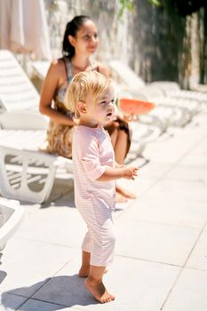 Little girl stands next to her mother sitting on a sun lounger with a watermelon in her hands. High quality photo