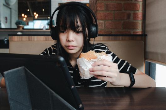 Girl eating burger watch your tablet and use headphones to listen to music or use social media.