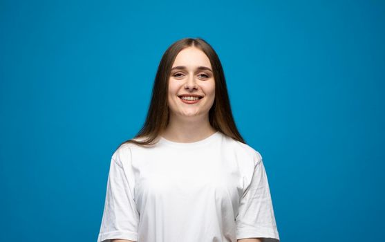 Happy, smiley and positive brunette girl in a white t-shirt looking at camera, smiling carefree on blue background studio. Emotions