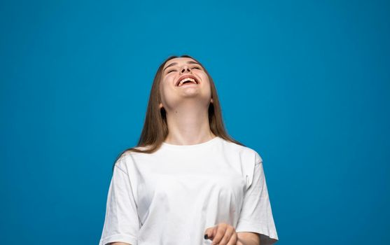 Young beautiful cute cheerful brunette girl in white t-shirt smiling looking at camera and showing a tongue over blue background studio. Emotions