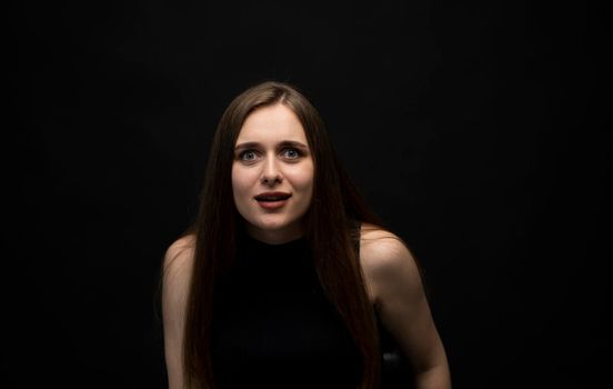 Portrait of upset young beautifil brunette girl with long hair in a black t-shirt in a black room. Emotion of angry and upset