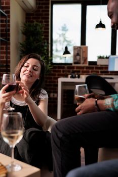 Multiethnic group of friends sitting in living room while drinking alcoholic beverages and talking together. Diverse happy people at home enjoying wine party while celebrating birthday.