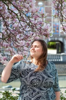 A plump, confident woman stands under a blooming magnolia and smiles. young millennial woman with brown curly hair smiling and waiting for a friend for a date. A full girl enjoys flowering and spring.