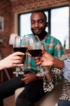 Joyful multiracial group of best friends toasting wineglasses while enjoying wine party at home. Happy smiling diverse people clinking glasses while celebrating birthday event.