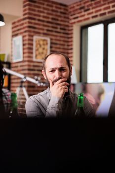 Worried caucasian man sitting at home while watching tragic news event online video on laptop. Anxious person sitting in living room while watching suspense movies on modern portable computer.