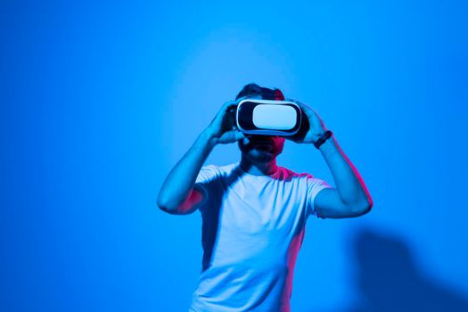 Closeup of young man in white t-shirt wearing virtual reality goggles in modern studio with a neon light