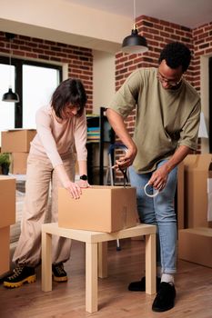 Diverse man and woman using sticky tape to pack things in boxes, moving together in new rented apartment flat. Relocating household and shipping cargo packages to property, life event.
