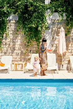 Mom with a watermelon in her hand leads a little girl to a sun lounger by the pool. High quality photo