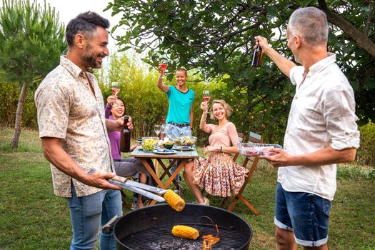 Friends making a toast at a barbecue dinner party while men are cooking. Weekend activities concept.