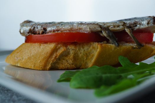 tapa of sardines on a slice of bread with tomato and olives on a white plate with a typical spanish white background