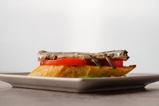tapa of sardines on a slice of bread with tomato and olives on a white plate with a typical spanish white background