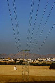 electric tower with cables, in the background a white village with cereal fields.