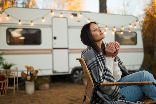 Beautiful caucasian woman is sitting wrapped in a plaid and drinking a warming drink outdoors. Traveling in a motor home in autumn