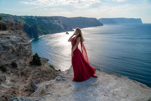 A girl with flowing hair in a long red dress stands on a rock above the sea. The stone can be seen in the sea. Sunny path to the sea from the sun