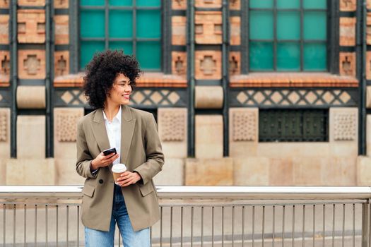 successful business woman smiles happy at city with a mobile phone in one hand and a coffee in the other, concept of urban lifestyle and technology, copy space for text