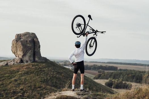 Professional Male Cyclist in Helmet Raising Hand with Bicycle while Standing on Top of Hill, Giant Stone, Miracle of Nature on the Background