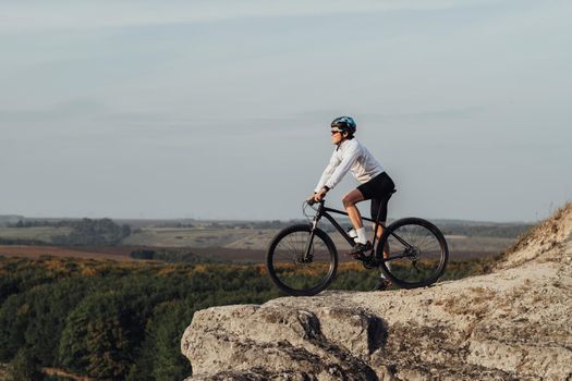 Equipped Professional Male Cyclist Standing with Mountain Bike on Edge of Rock and Enjoys Panoramic View Outdoors