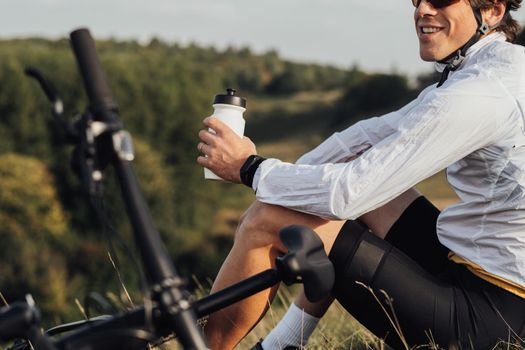 Professional Male Cyclist Drinking Water from Bottle, Man Sitting Near Bicycle During His Journey Outdoors in Countryside
