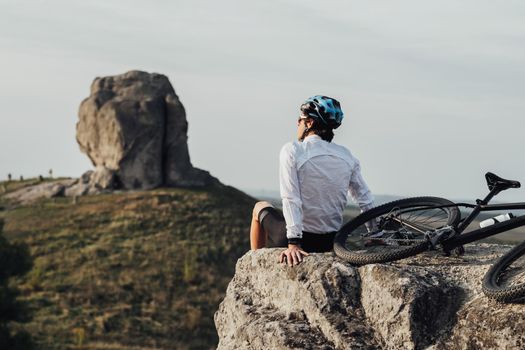 Equipped Professional Male Cyclist Sitting with Mountain Bike on Edge of Rock and Enjoys Panoramic View on Giant Stone, Miracle of Nature
