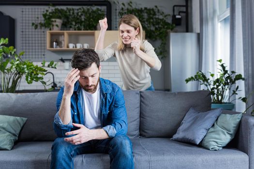 Young family, couple at home, man and woman quarrel, conflict, woman shouting and waving hands. Man sitting on sofa, holding head, listening