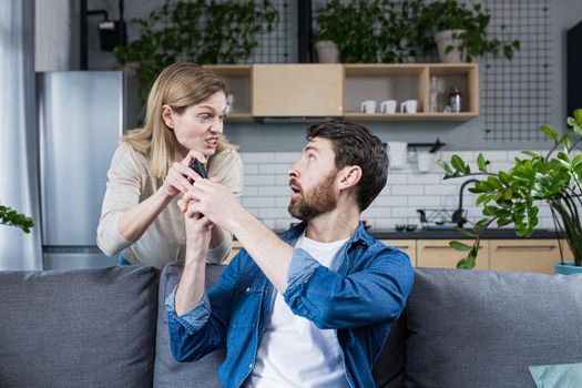 Conflict in the family. An angry, annoyed woman's wife takes the phone from the man who is sitting on the couch at home and constantly uses the phone.