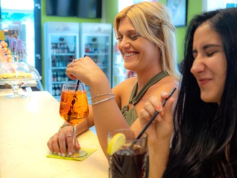 caucasian blond woman drinking spritz close to a latino woman drinking cola drink at the pub.