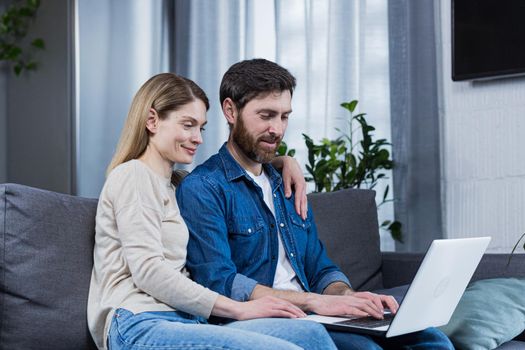 Happy young married couple man and woman together at home, sitting on sofa smiling and using laptop