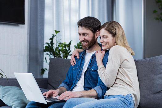 Happy young married couple man and woman together at home, sitting on sofa smiling and using laptop