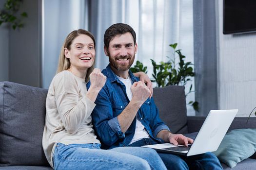 Young couple, family, husband and wife sitting on sofa at home, using laptop, winning something, getting good news, rejoicing, smiling, hugging