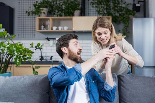 Conflict in the family. An angry, annoyed woman's wife takes the phone from the man who is sitting on the couch at home and constantly uses the phone.