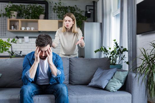 Young family, couple at home, man and woman quarrel, conflict, woman shouting and waving hands. Man sitting on sofa, holding head, listening