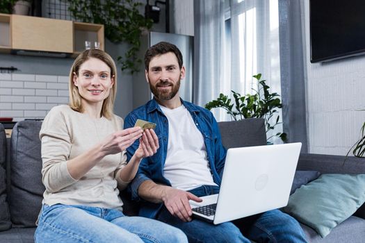 Happy married couple man and woman sitting at home on the couch using laptop for online shopping, holding a credit bank card