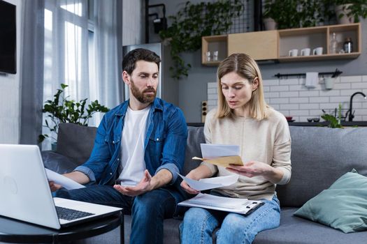 Young family, husband and wife, reviewing their bills, loan and mortgage agreements, sitting on the couch at home, working with documents on a laptop, embarrassed, upset
