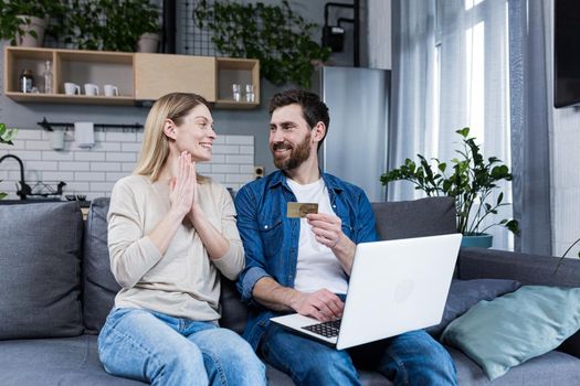 Happy married couple man and woman sitting at home on the couch using laptop for online shopping, holding a credit bank card