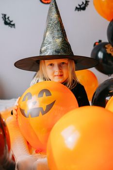 Children's Halloween - a girl in a witch hat and a carnival costume with airy orange and black balloons at home. Ready to celebrate Halloween.