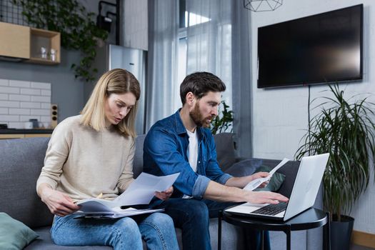 Young family, husband and wife, reviewing their bills, loan and mortgage agreements, sitting on the couch at home, working with documents on a laptop, embarrassed, upset