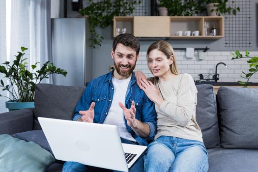 Young couple, family, man and woman sitting at home on the couch talking on video call from laptop to family, friends. They wave, smile, greet