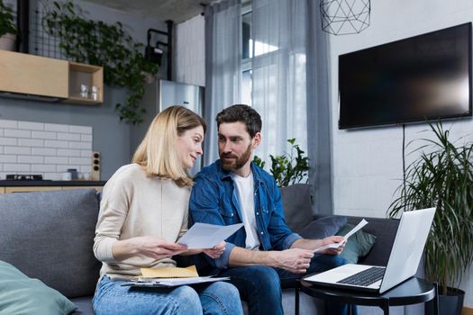 Young family, husband and wife, reviewing their bills, loan and mortgage agreements, sitting on the couch at home, working with documents on a laptop, embarrassed, upset