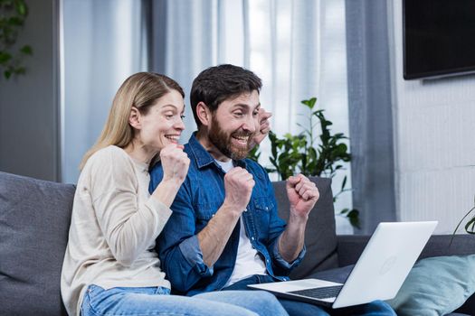 Young couple, family, husband and wife sitting on sofa at home, using laptop, winning something, getting good news, rejoicing, smiling, hugging