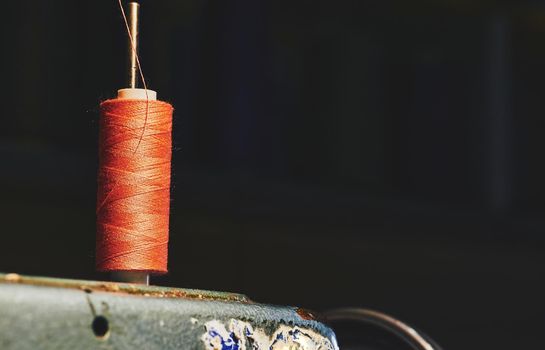 a long, thin strand of cotton, nylon, or other fibers used in sewing or weaving. Orange spool of thread on an old gray sewing machine and a dark background.