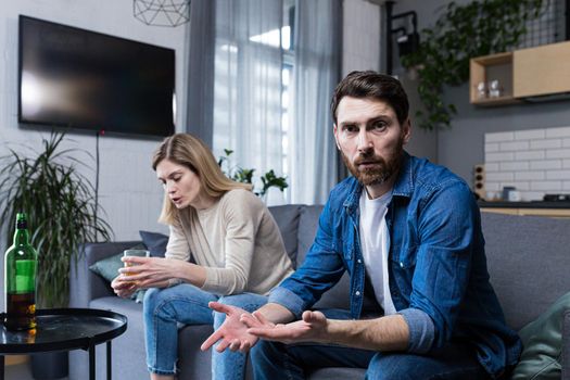 Female alcoholism, husband in despair, woman wife drinking hard alcohol, couple sitting on sofa in living room