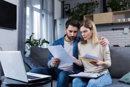 Young family, husband and wife, reviewing their bills, loan and mortgage agreements, sitting on the couch at home, working with documents on a laptop, embarrassed, upset