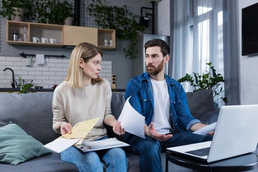 Young family, husband and wife, reviewing their bills, loan and mortgage agreements, sitting on the couch at home, working with documents on a laptop, embarrassed, upset