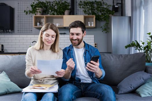A man and a woman are happy at home, make online payments, using the phone and banking application, sitting on the couch and holding bills